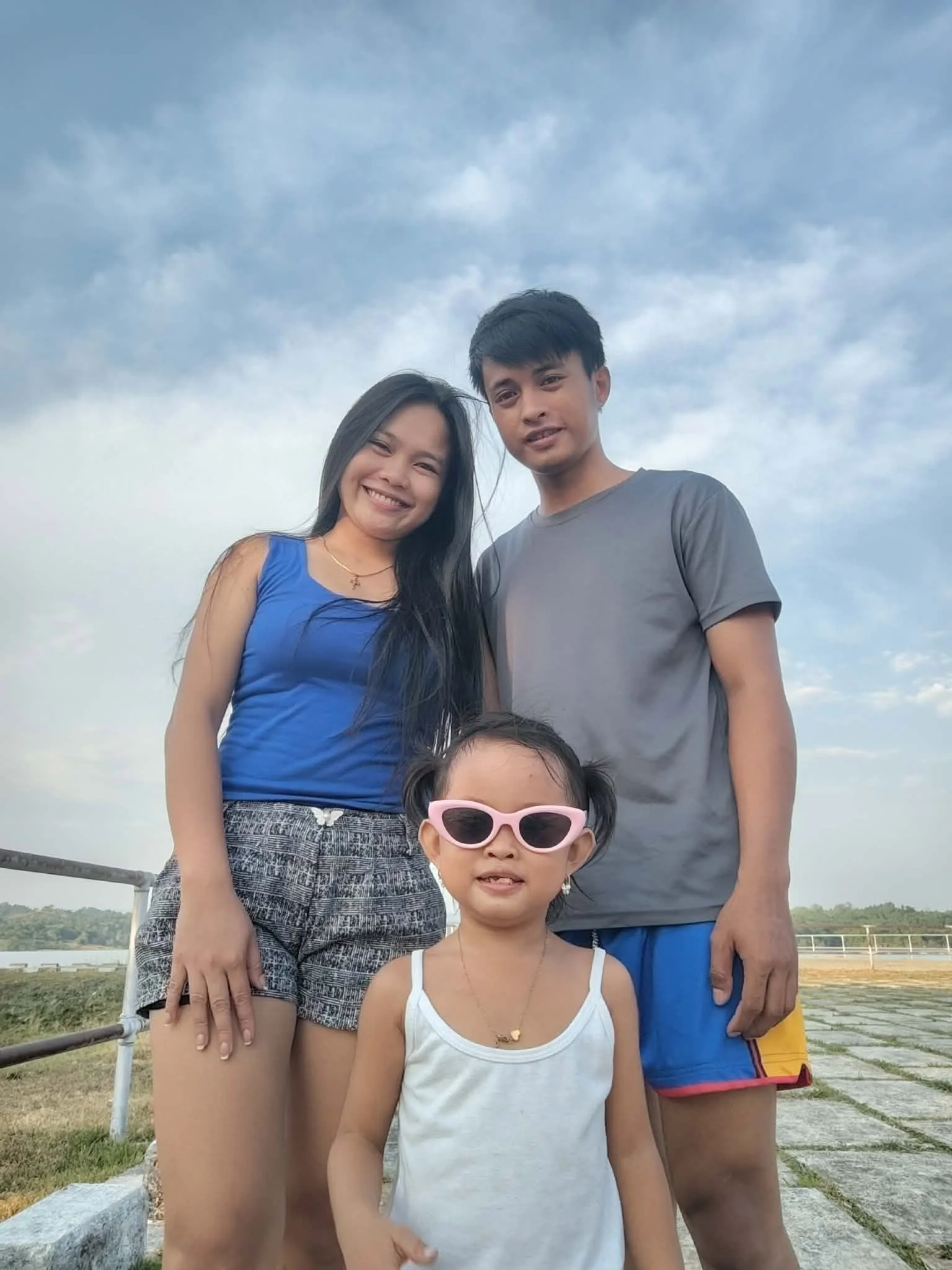 A family of four, including a woman in a blue top, a man in a gray T-shirt, a young girl with pink sunglasses, and a girl with necklace, standing outdoors with a cloudy sky and a body of water in the background.