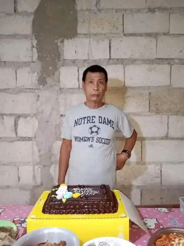 A man stands behind a table with a birthday cake on it. The man is wearing a white T-shirt with "Notre Dame Women's Soccer" written on it. The background is a rough concrete wall.
