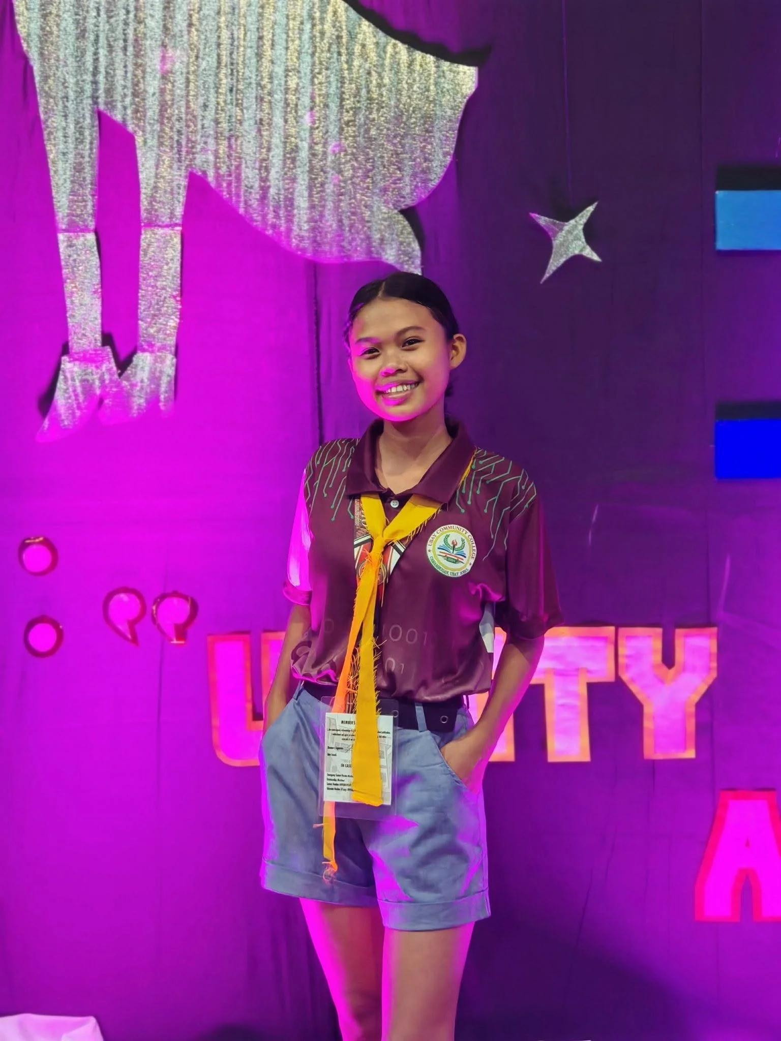 A young woman in a brown uniform shirt with yellow neckerchief standing in front of a purple background with stars and geometric shapes.