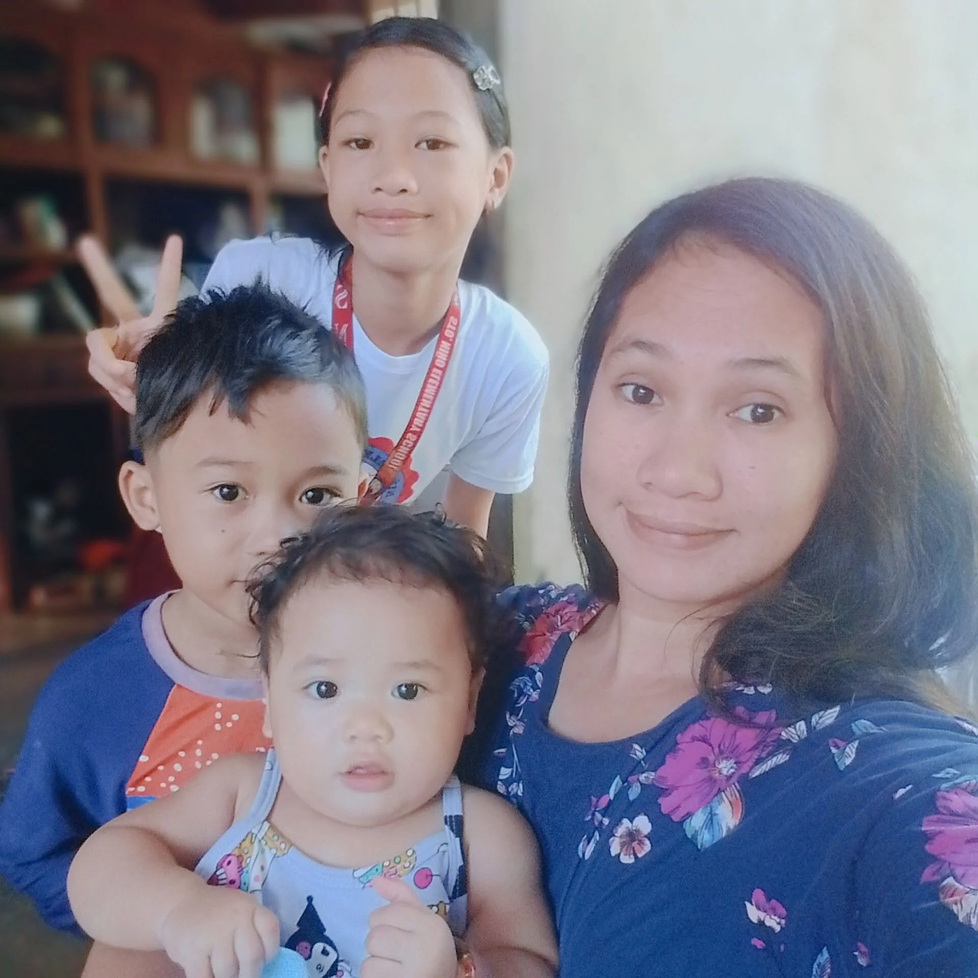 Three young children and a woman are taking a selfie together. The children have dark hair and are wearing colorful clothes, and the woman has long dark hair and is smiling. One child is making a peace sign.