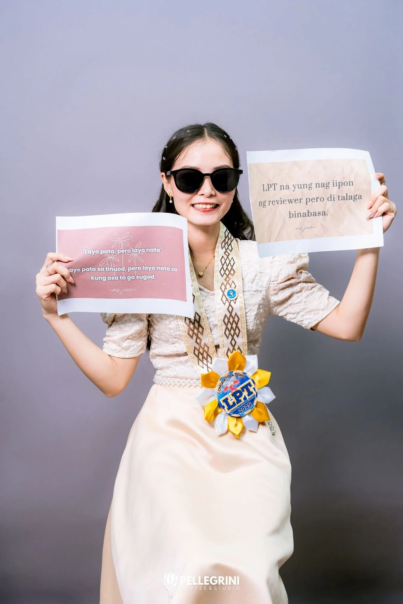 A woman wearing black sunglasses and earrings, smiling, dressed in traditional Filipino attire, holding two signs with Tagalog text, wearing a medal with the abbreviation 'LPT'.