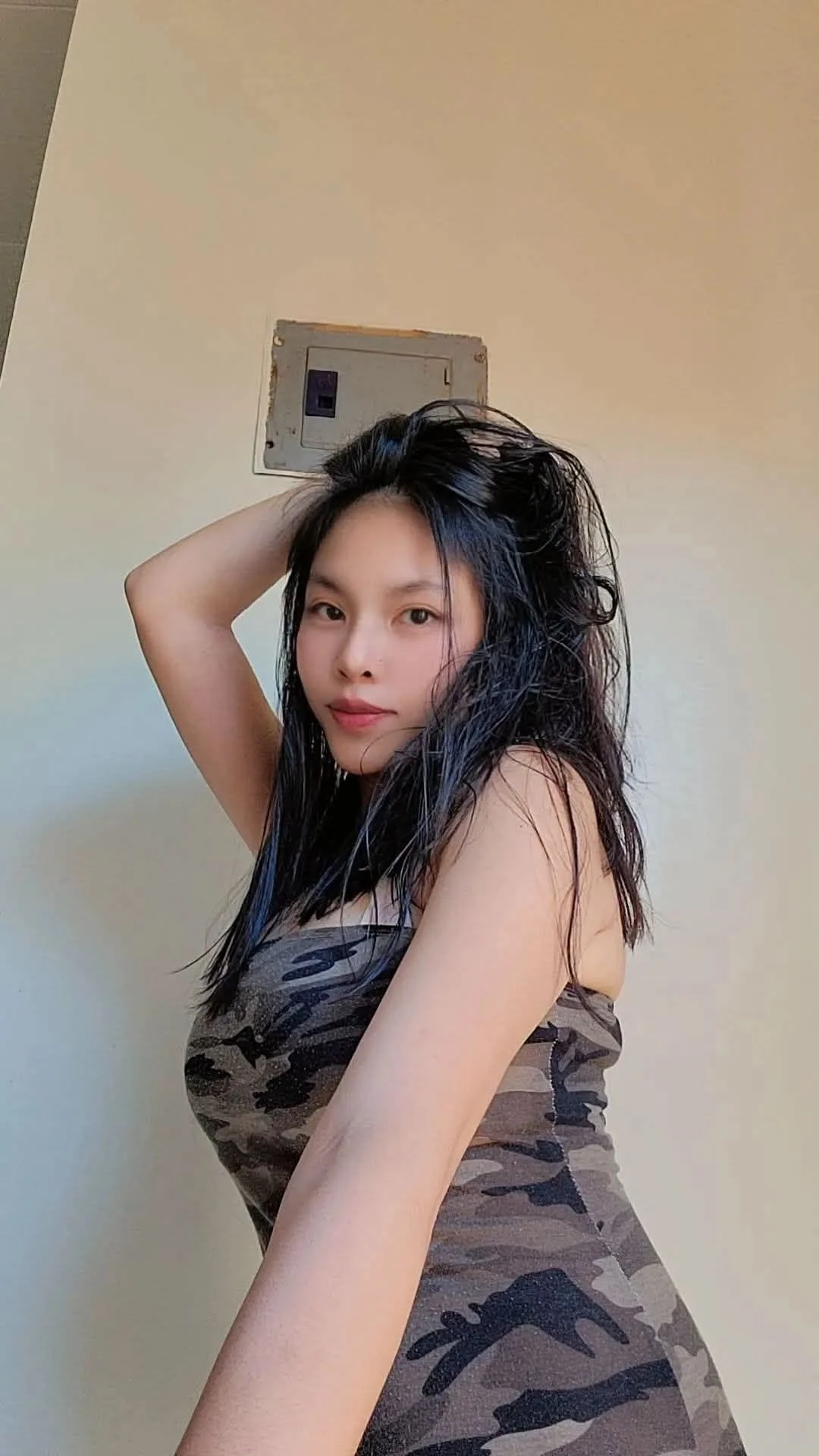 A woman with long, wet black hair wearing a camouflage tank top, posing with one hand in her hair in front of a beige wall.