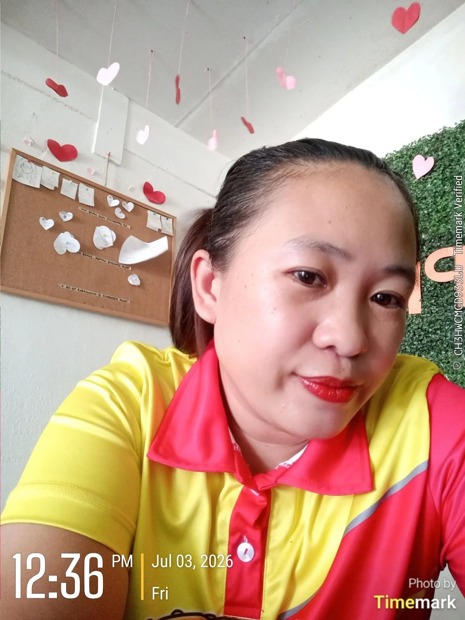 Close-up of a woman with dark hair pulled back, wearing a bright yellow and red uniform, smiling slightly. A cork bulletin board with white and red paper hearts and notes is visible in the background. The image includes a timestamp showing 12:36 PM o