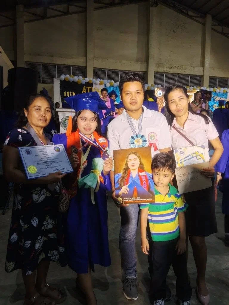 Group of five people, including a young girl in graduation gown and cap, celebrating graduation with medals, certificate, and photo, at a school event with balloons and banners in the background.