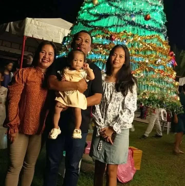 Family of four, including a toddler girl, standing in front of a decorated Christmas tree outdoors at night.
