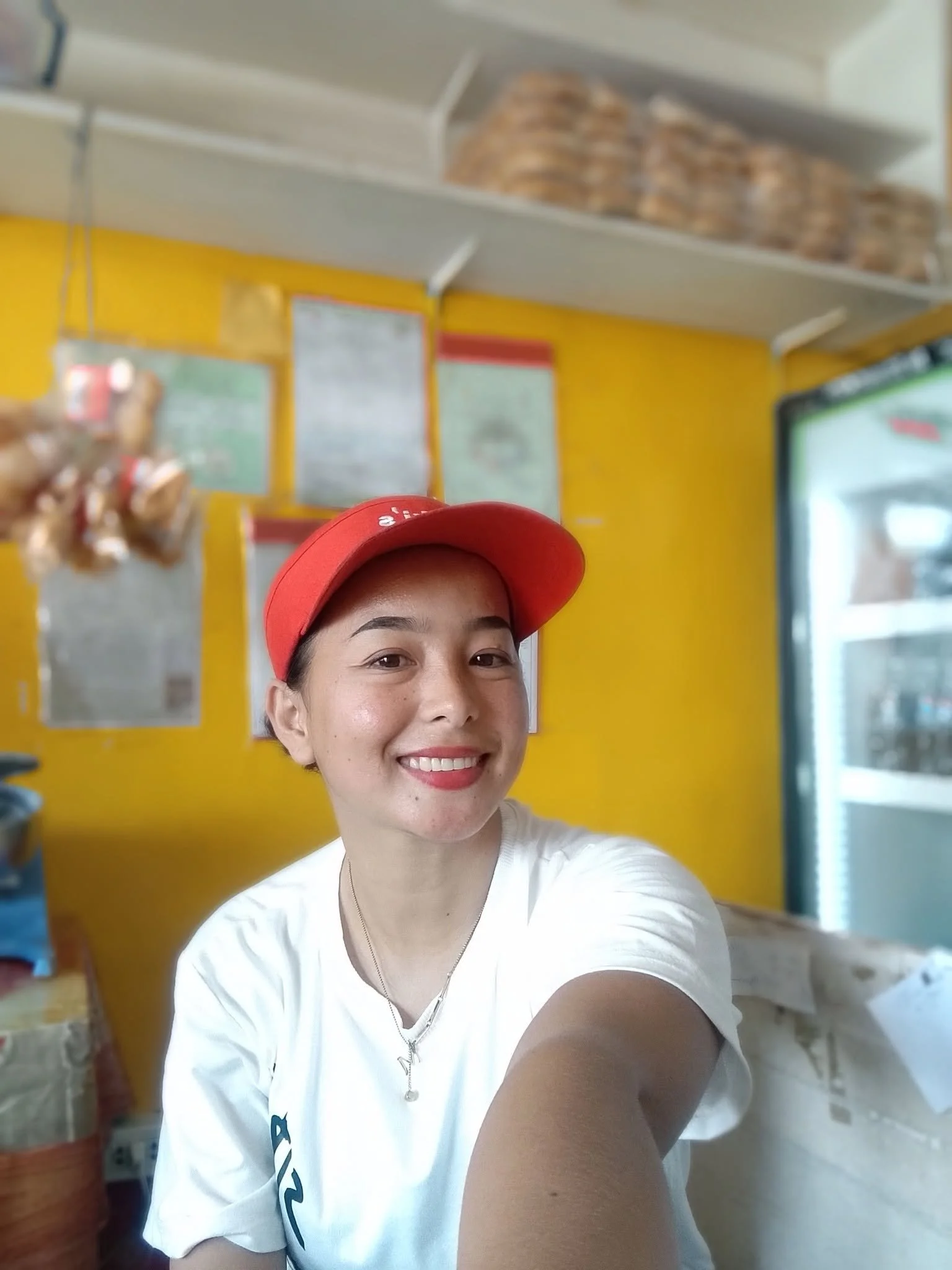 A woman wearing a red baseball cap and white shirt smiling while taking a selfie inside a store with yellow walls and a glass display case.