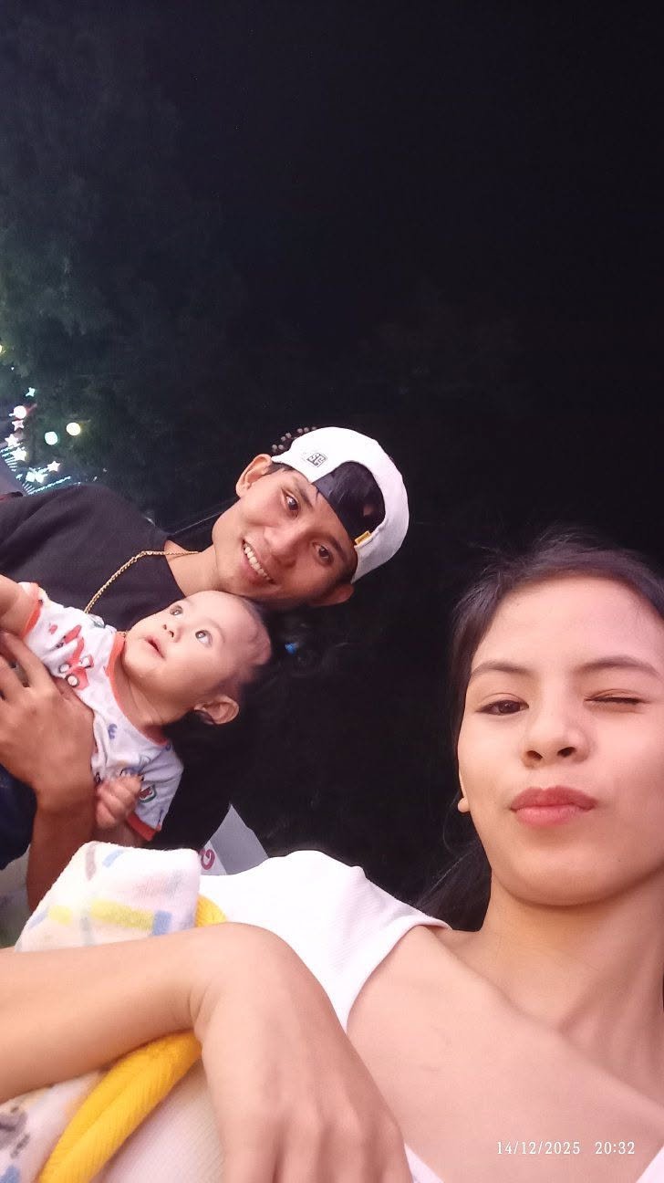 Three people, two adults and one baby, taking a selfie at night outdoors. The woman on the right is winking, the man on the left is smiling, and the baby in the middle is looking up with a curious expression. Background shows dark night sky and some 