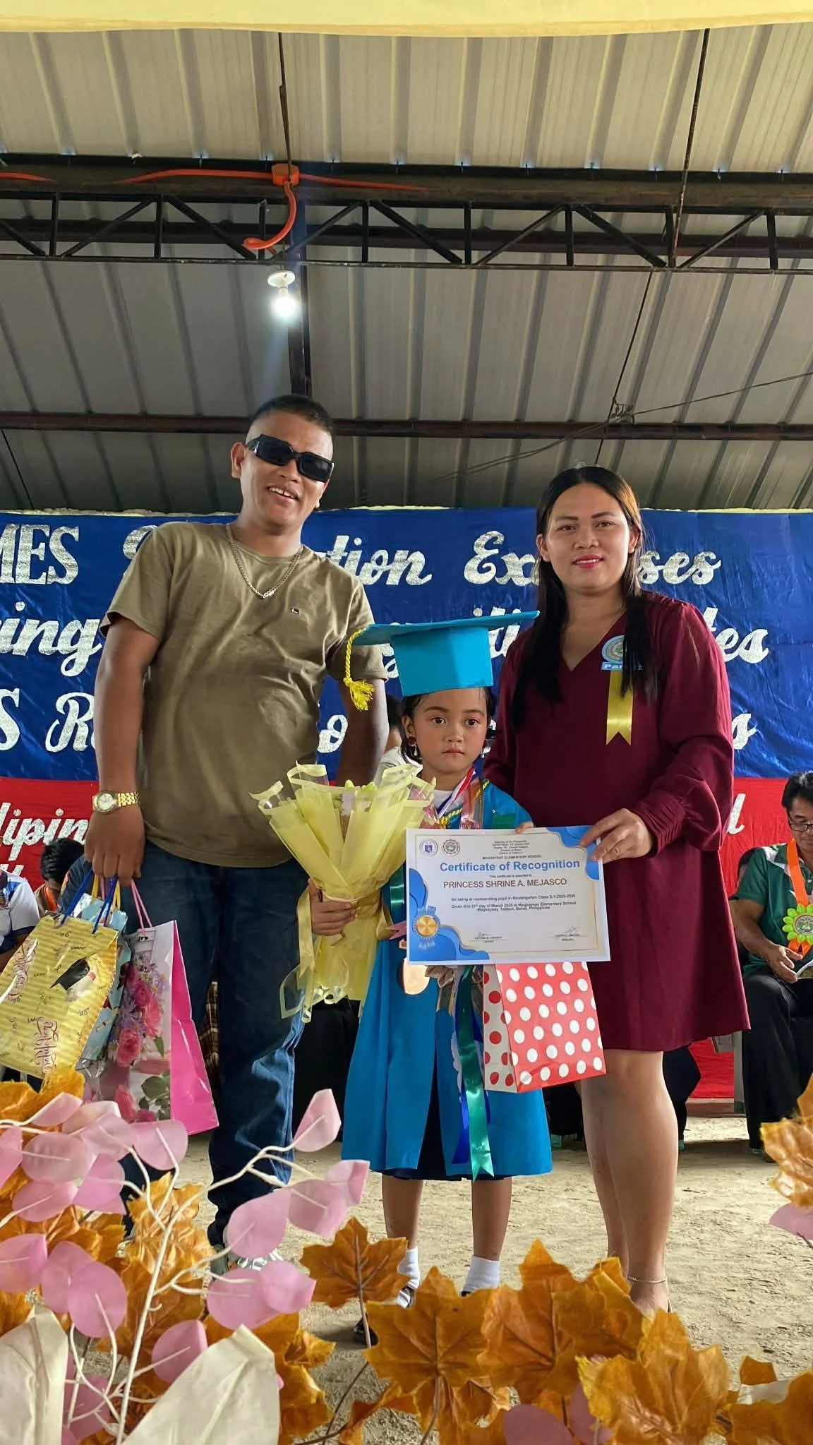 A young girl in a blue graduation gown and cap holding a certificate of recognition, flanked by two adults, standing under a covered area during a graduation or recognition ceremony. The girl also has a bouquet of flowers, and there are gift bags in 