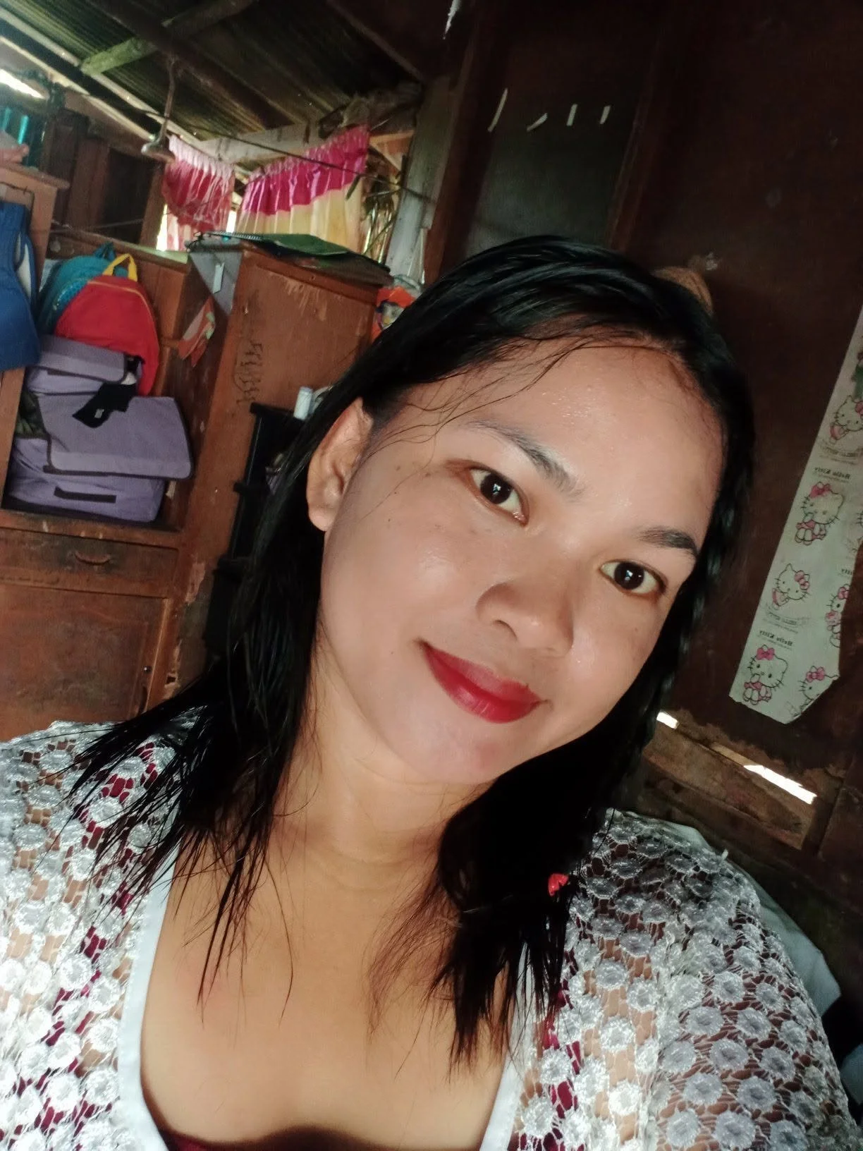 A woman with black, wet hair, wearing red lipstick and a white lace top, smiling inside a wooden house with luggage, bags, and Hello Kitty curtains in the background.