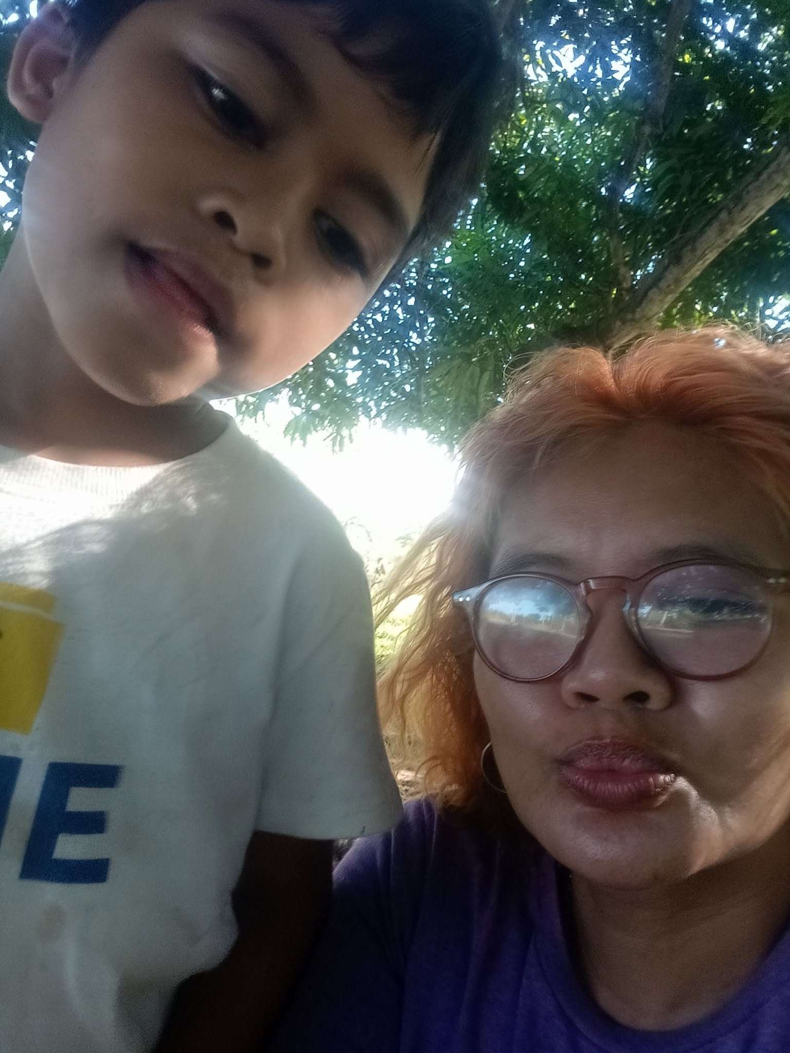 A close-up selfie of a child with short dark hair and a woman with red hair and glasses, both outdoors under a tree with green leaves.