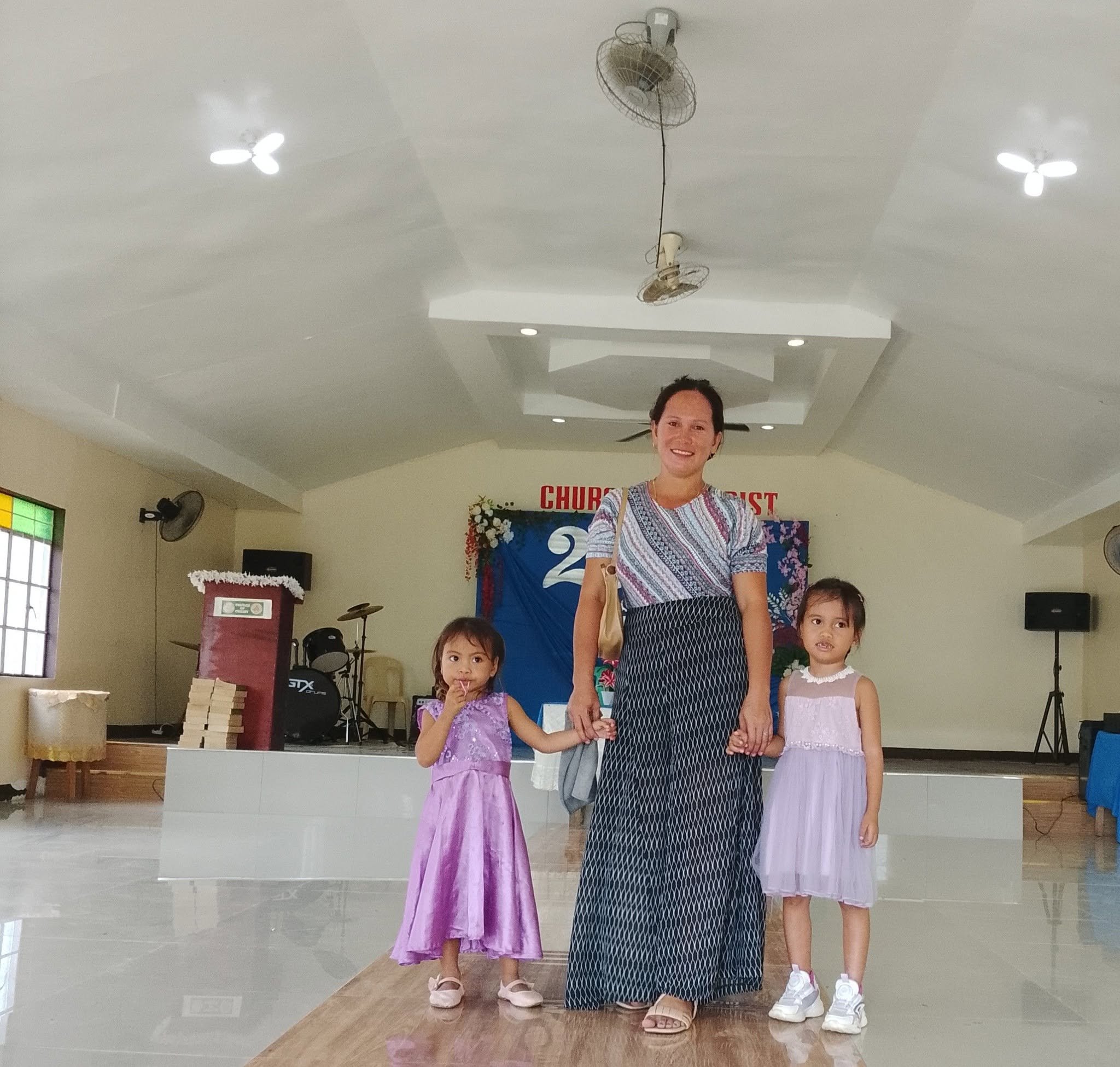 A woman and three young girls standing on a stage indoors. The woman is smiling, wearing a patterned dress, and holding hands with two girls. The girls are wearing pink dresses. In the background, there is a blue banner with the word 'CHURCH' on it, 