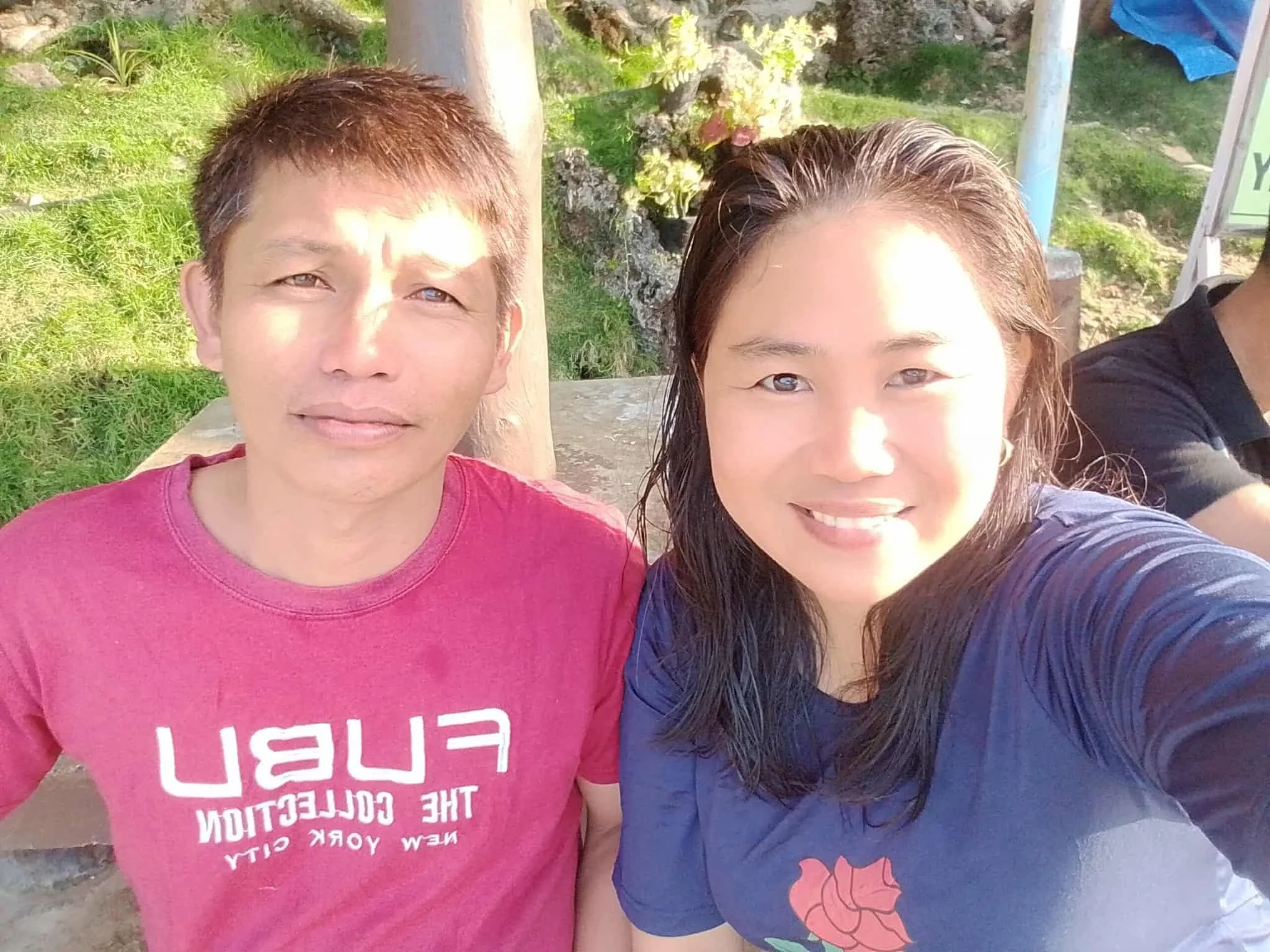 Close-up of two people taking a selfie outdoors with a green grassy area and plants in the background. One is wearing a pink shirt and the other a blue shirt with a floral design.