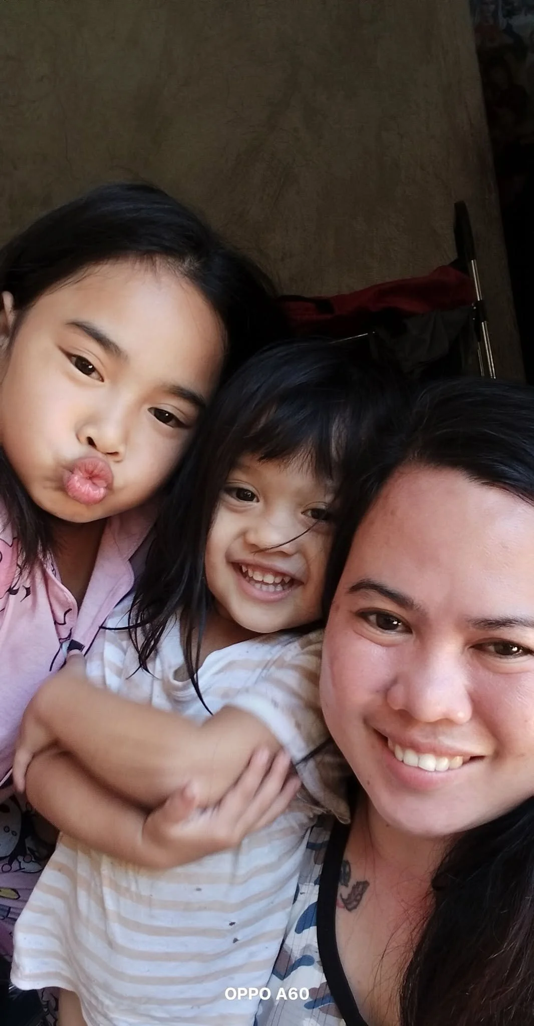 A smiling woman takes a selfie with two young girls, one making a duck face and the other smiling openly, all close together indoors.