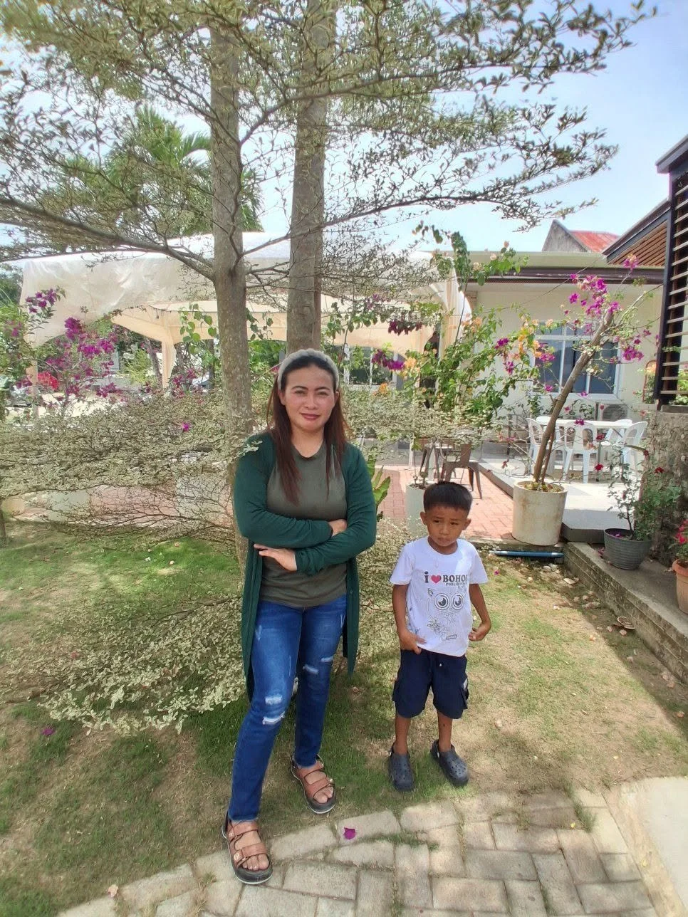 A woman with brown hair in casual clothing standing outdoors in a garden with a young boy who is wearing a white t-shirt with a design, dark shorts, and Crocs, in front of trees, plants, and potted flowers, during daytime.
