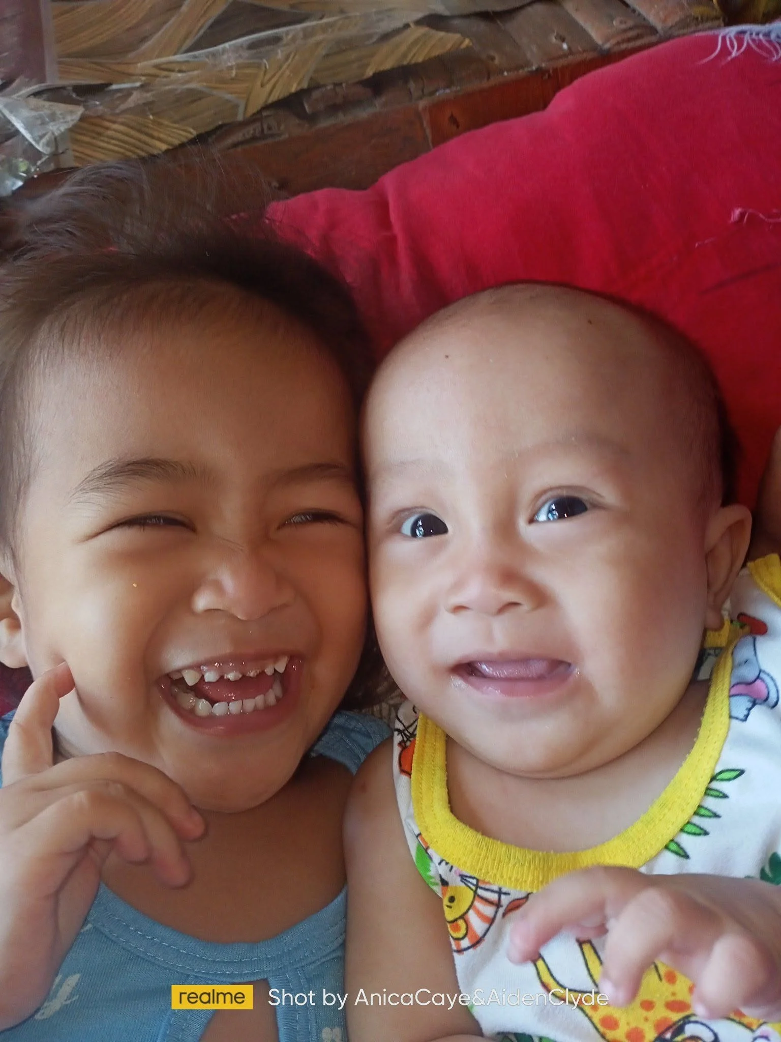 Two young children lying on the floor, smiling and looking up, close together, with a brick wall in the background.