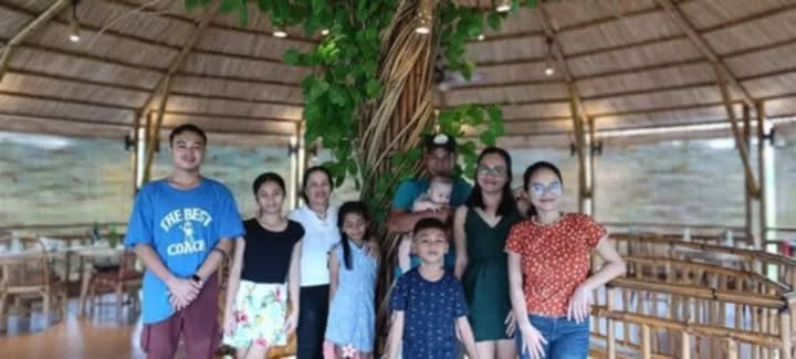 Group of eight children and two adults standing inside a bamboo structure with a large tree in the background.