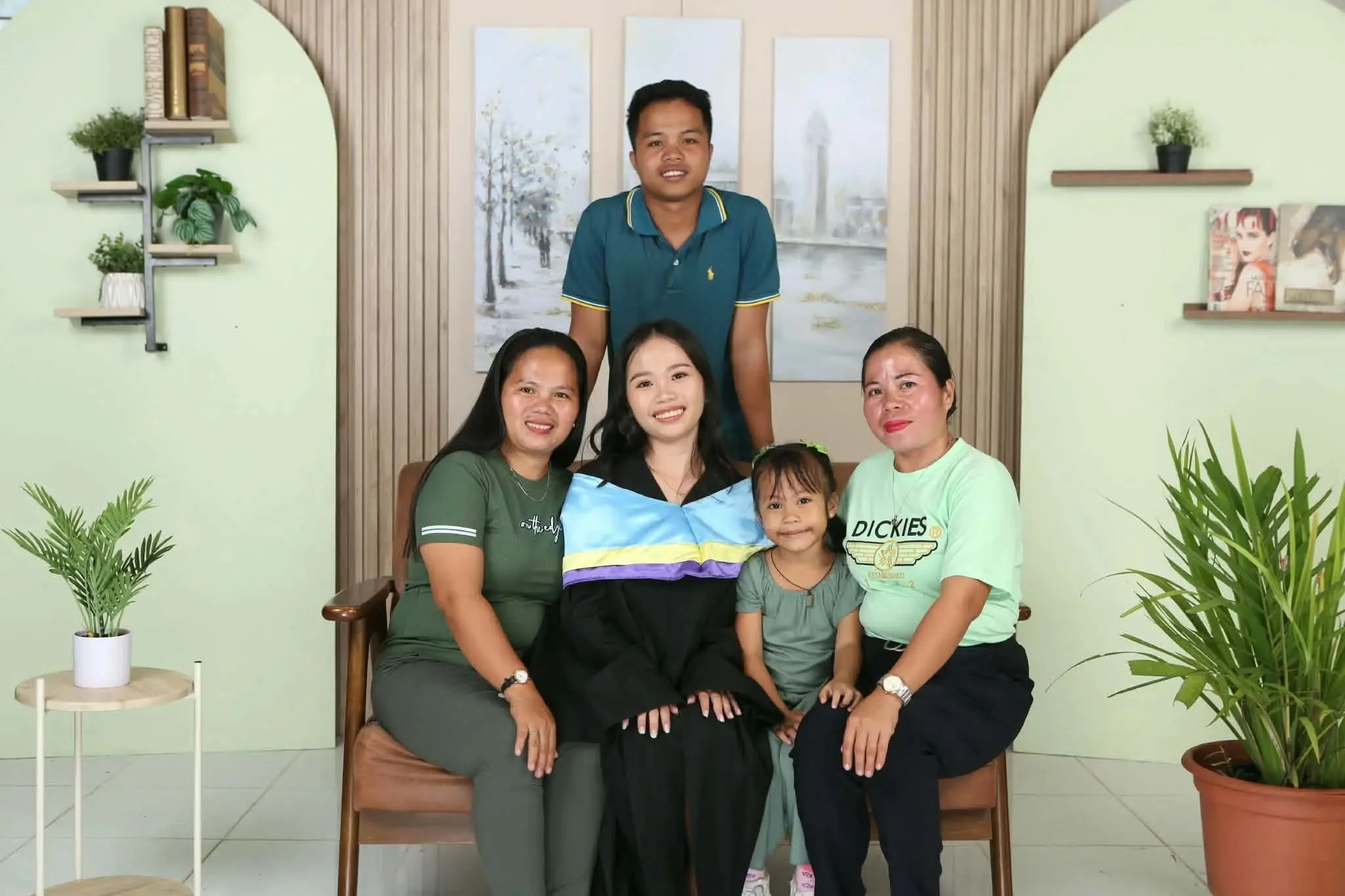 A group of five people, three women, one man, and a young girl, sitting and standing in a room with light green walls and potted plants, smiling for a photo.