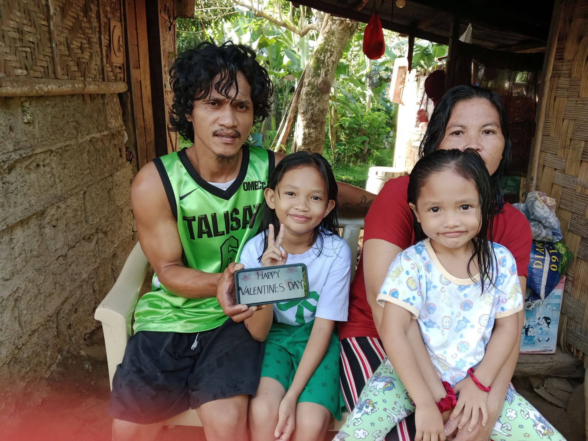 A group of four people, including two children, sitting outdoors in a rustic setting with trees and wooden structures. One child is holding a small sign that reads "HAPPY VALENTINES DAY."