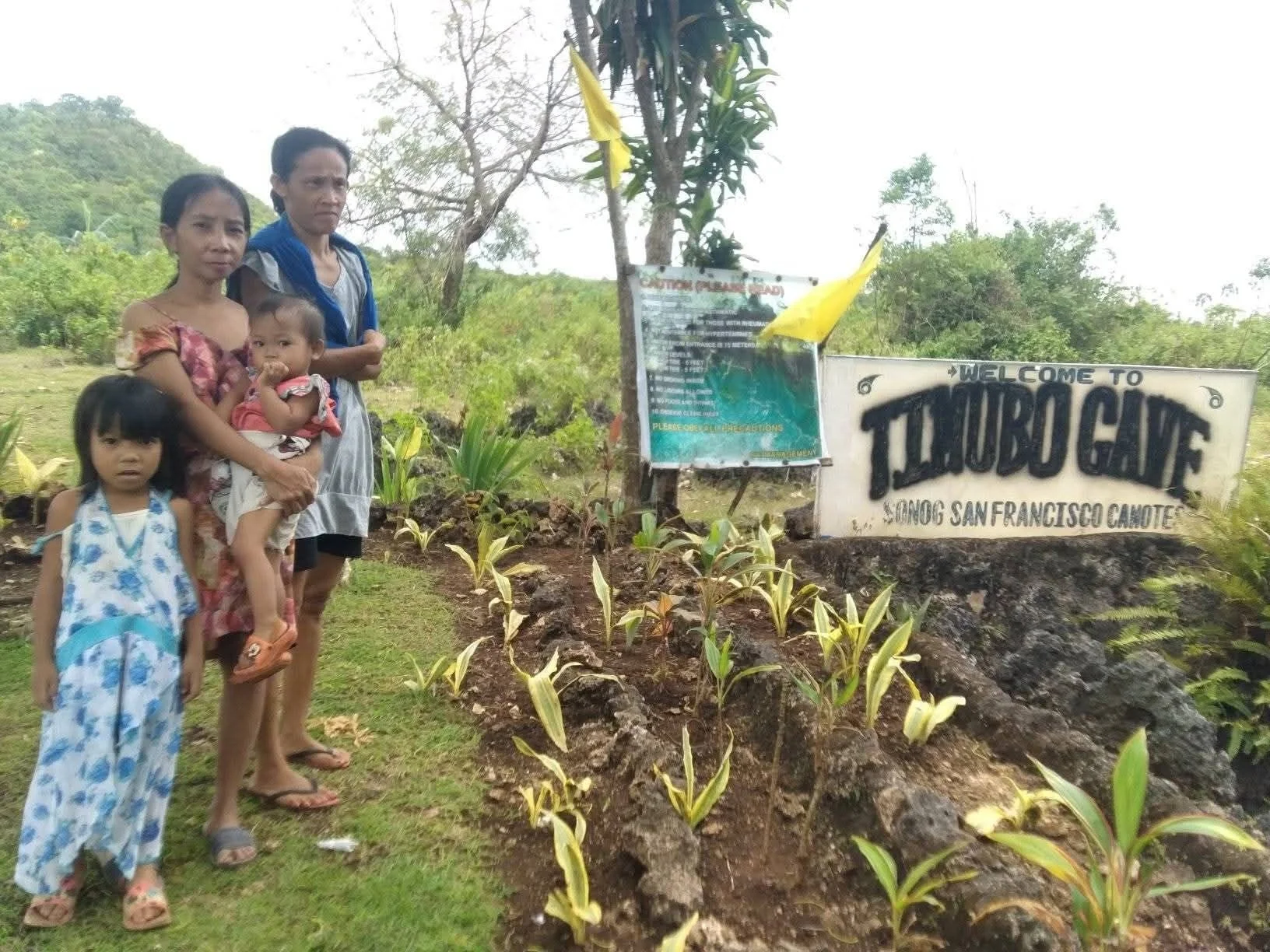 A family of four standing outdoors near a sign that says "Welcome to Tuluba Gaye" in Sebang San Francisco Camotes, with a landscaped garden and greenery in the background.