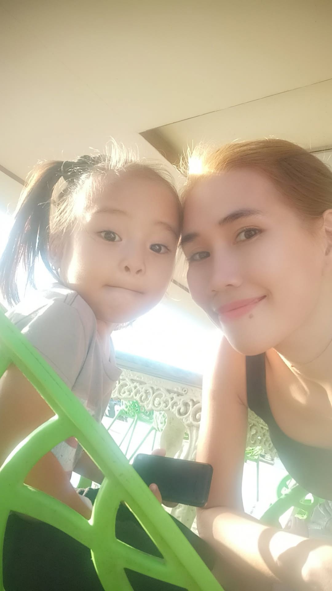 A woman and a young girl taking a selfie, both smiling, with sunlight shining behind them, outdoors on a sunny day.