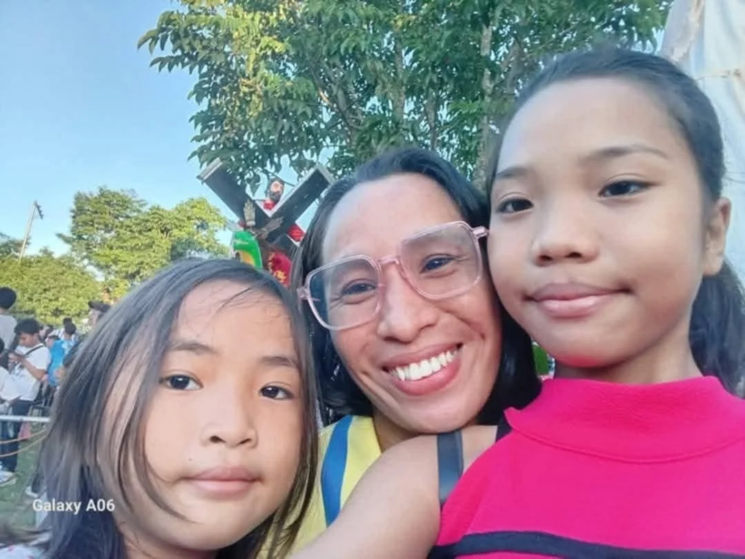 A woman smiling among two young girls, outdoors with trees and a crowd in the background, sunny day.