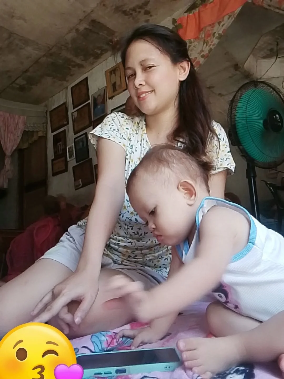 A woman and a young boy sitting together in a room with pictures on the wall, a fan in the background, and a phone on the bed. The woman is smiling with eyes half-closed, and the boy is focused on something off-camera.
