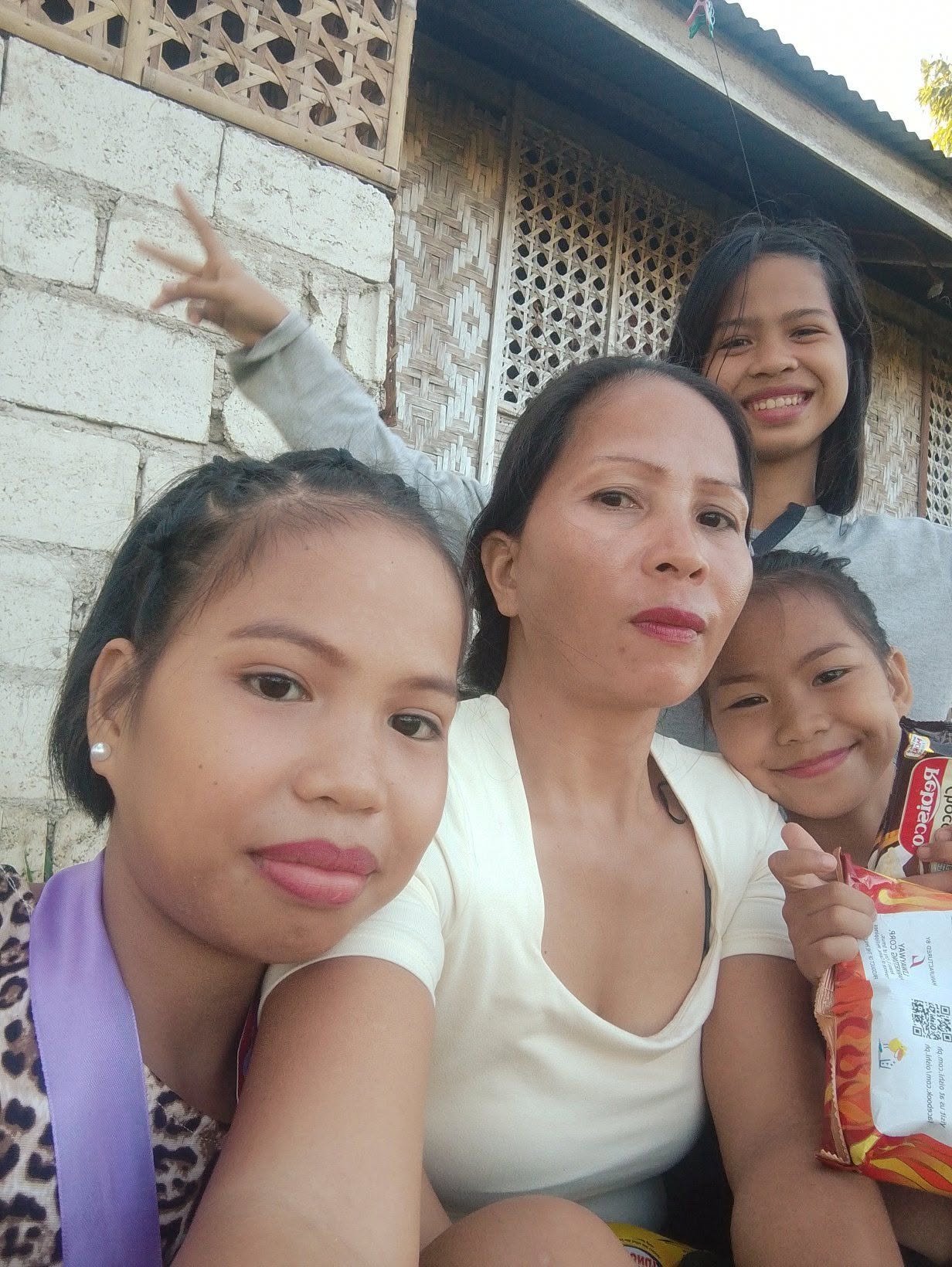 A woman with three girls posing for a selfie outdoors in front of a rustic building with bamboo and stone walls.