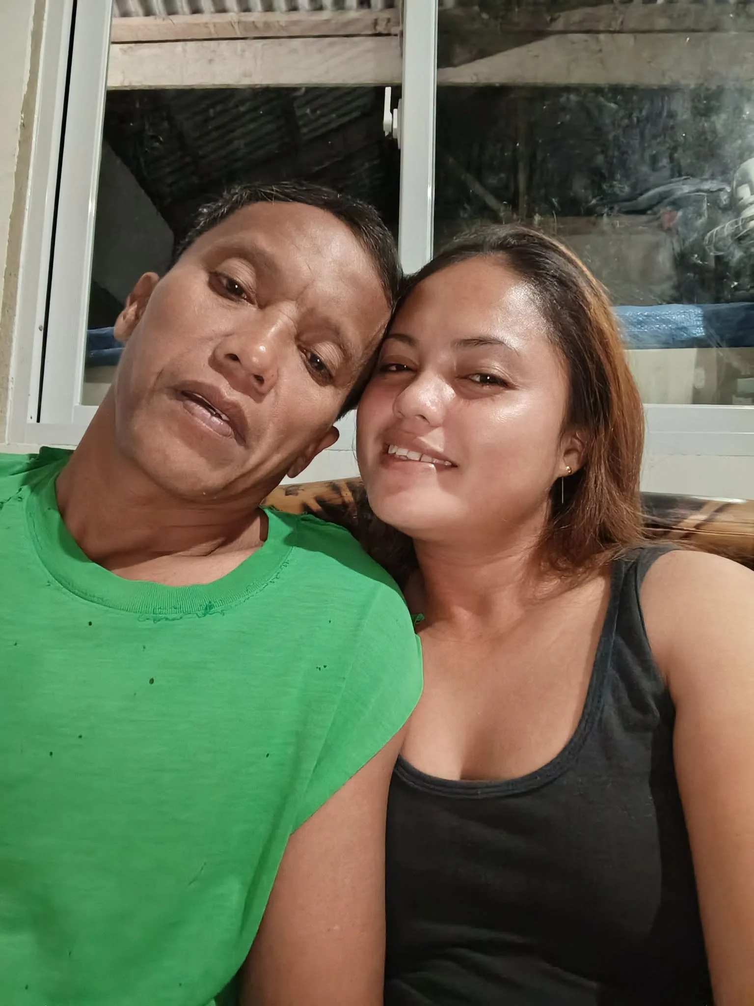 A middle-aged man and a young woman smiling closely for a selfie indoors in front of a window with a wooden roof visible outside.