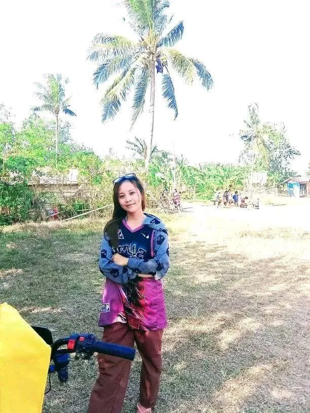 A girl standing outdoors in a field with palm trees in the background. She is wearing a jacket, pink shirt, and brown pants, with sunglasses on her head and long dark hair. There are other people sitting in the distance near some small structures.