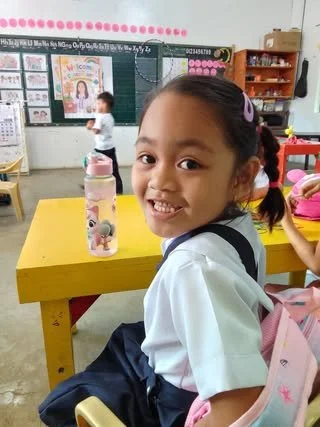 Young girl smiling in classroom with yellow desk, water bottle, and classroom decorations in the background.