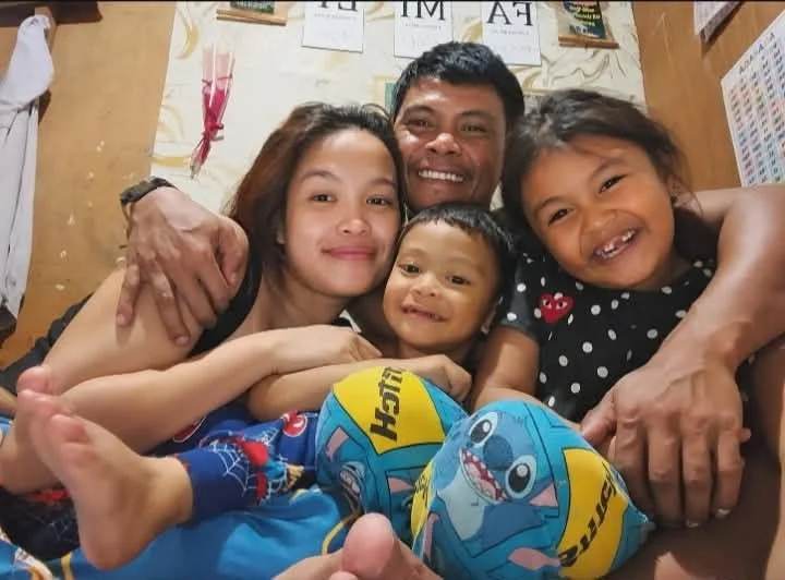 A family of five sitting close together, smiling, in a casual indoor setting with posters and papers on the wall behind them.