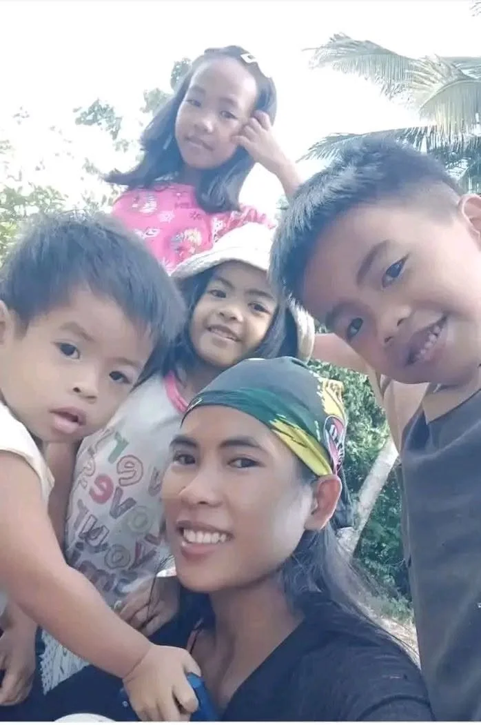 A woman with a bandana on her head taking a selfie with four children outdoors during daytime, with trees and palm leaves in the background.