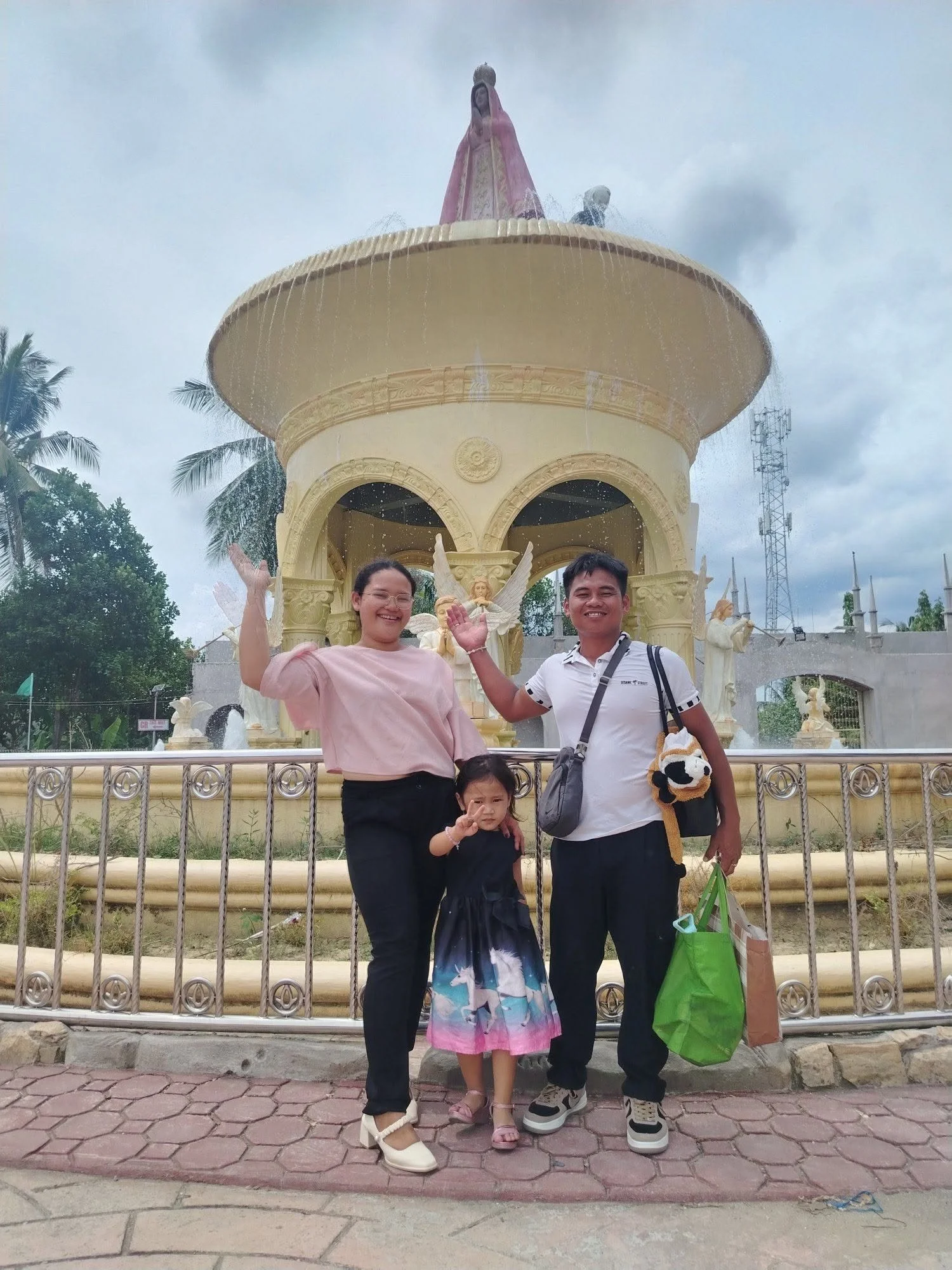 A group of four people standing in front of a decorative fountain with angel sculptures, posing for a photo outdoors on a cloudy day. The group includes a woman, a man, and two young girls.