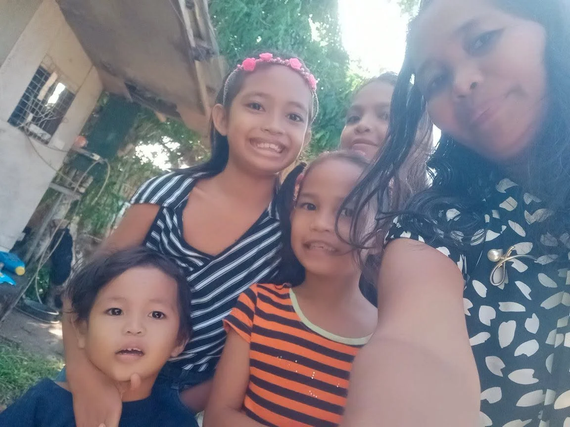Six people, including five children, posing outdoors for a selfie, smiling, with trees and a house in the background.