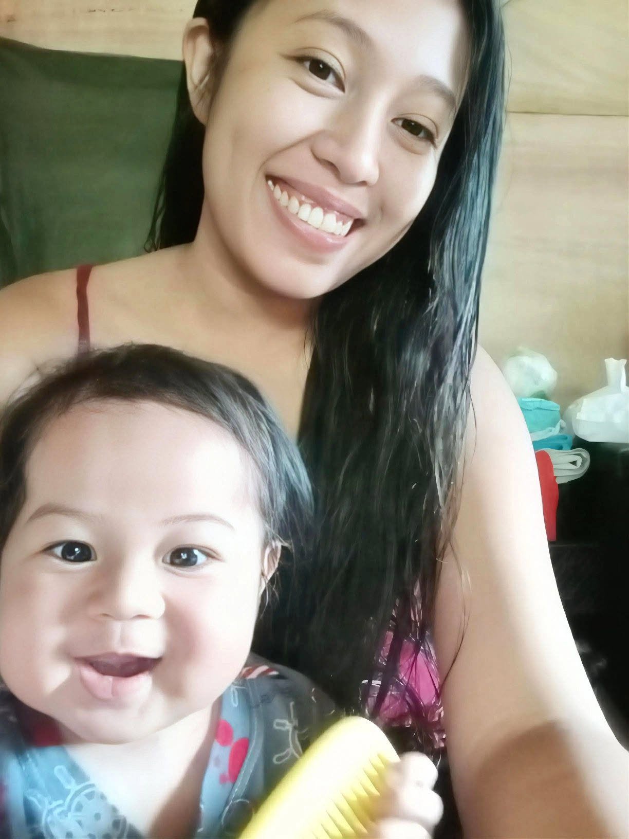 Smiling woman with long black hair holding a young girl with dark hair and bright eyes, in a room with a wooden wall and some clutter.