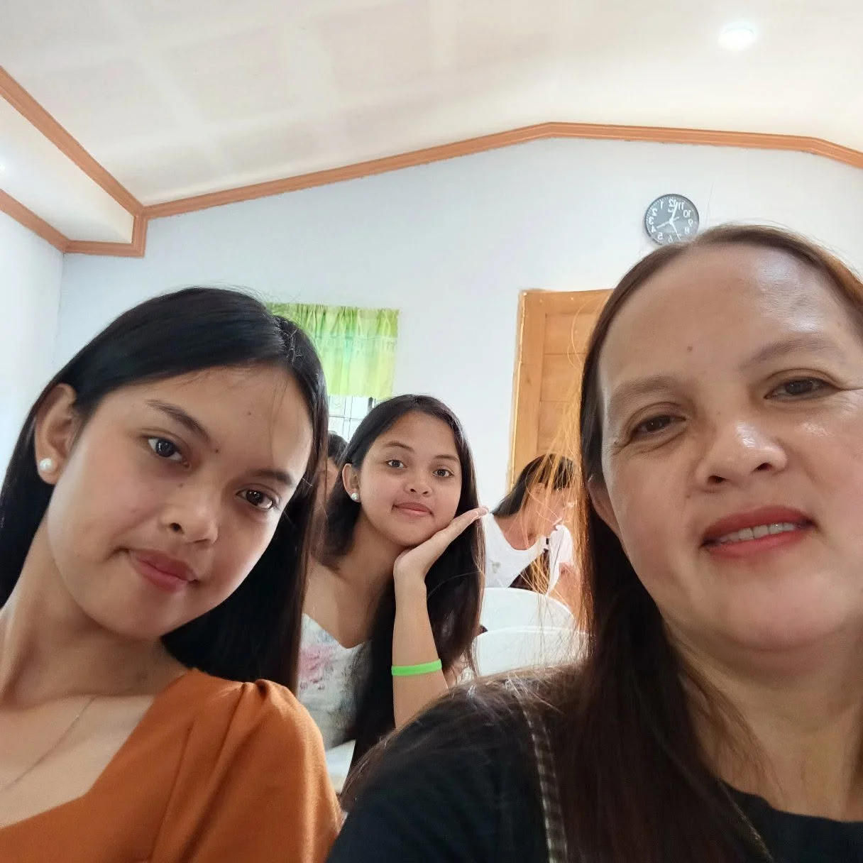 Group of four women taking a selfie inside a room with a white wall, wooden door, and clock, with one woman in the foreground, two women in the middle, and one woman in the background.