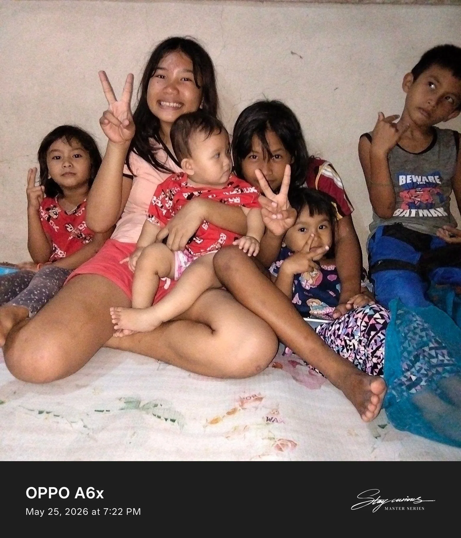 A group of children and a woman sitting on a bed against a plain wall. The children are making peace signs and appear to be relaxed and playful. The woman is smiling and holding one of the children. The scene is casual and cheerful.
