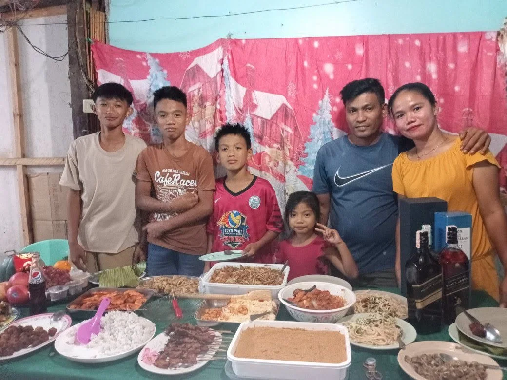 Family of seven posing behind a table filled with various dishes and beverages in a home setting.