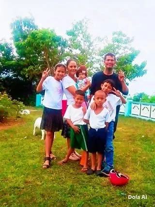 A group of seven people, including children and an adult male, standing outdoors on grass with trees in the background. They are smiling and posing for the photo.