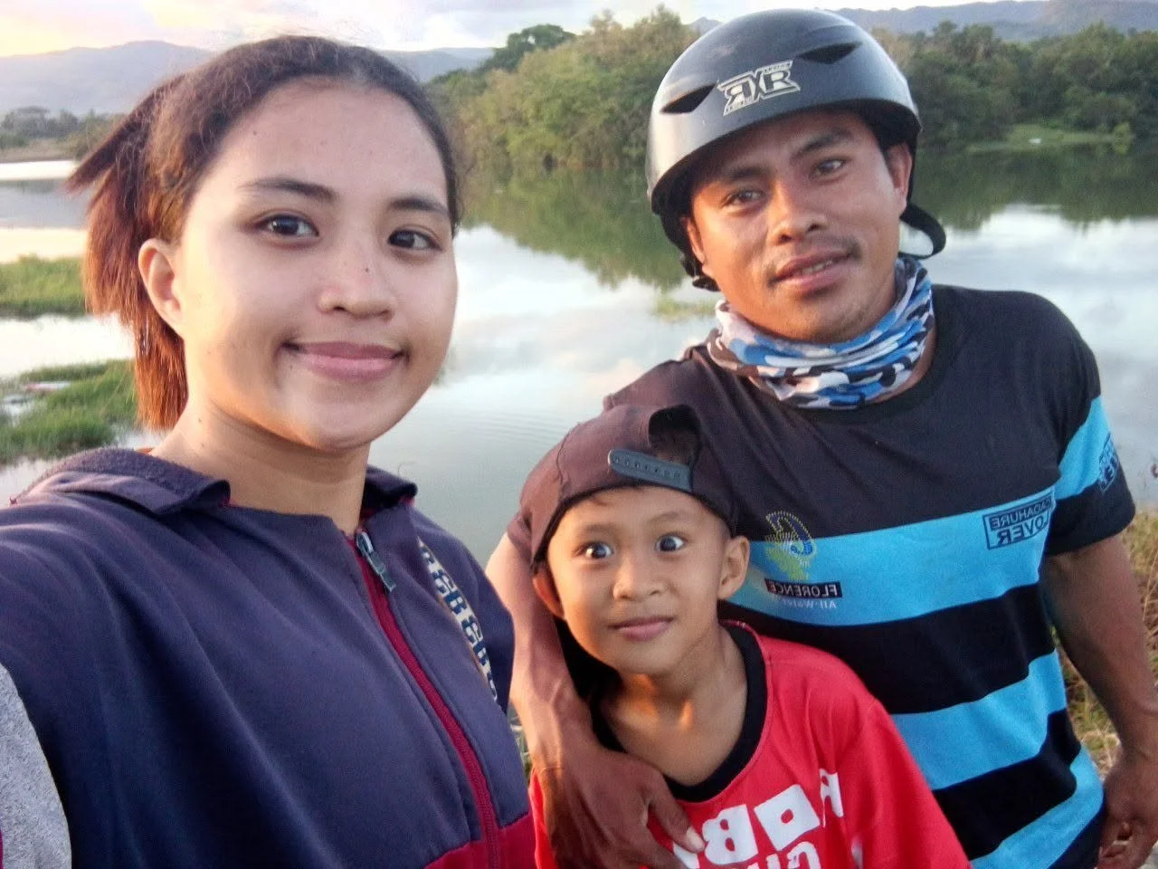 Three people outdoors near a body of water, smiling at the camera, with trees and mountains in the background. One woman, one man wearing a helmet, and a young boy.