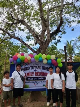 Group of five people standing outdoors in front of a sign with colorful balloons, trees, and palm trees behind them, celebrating a family reunion on May 24, 2026.