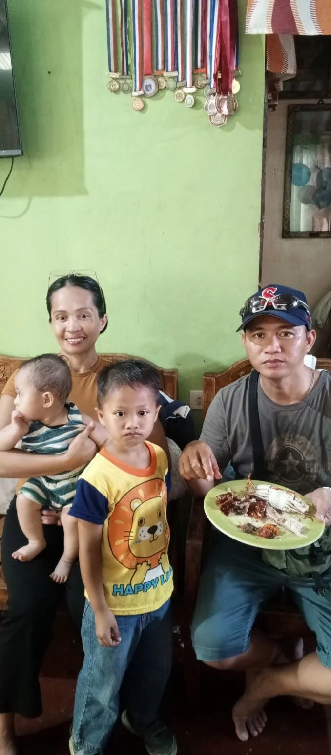 Family of four with a baby, sitting in a room with a green wall and a medal display, one person eating with a plate of food, two children looking at the camera.