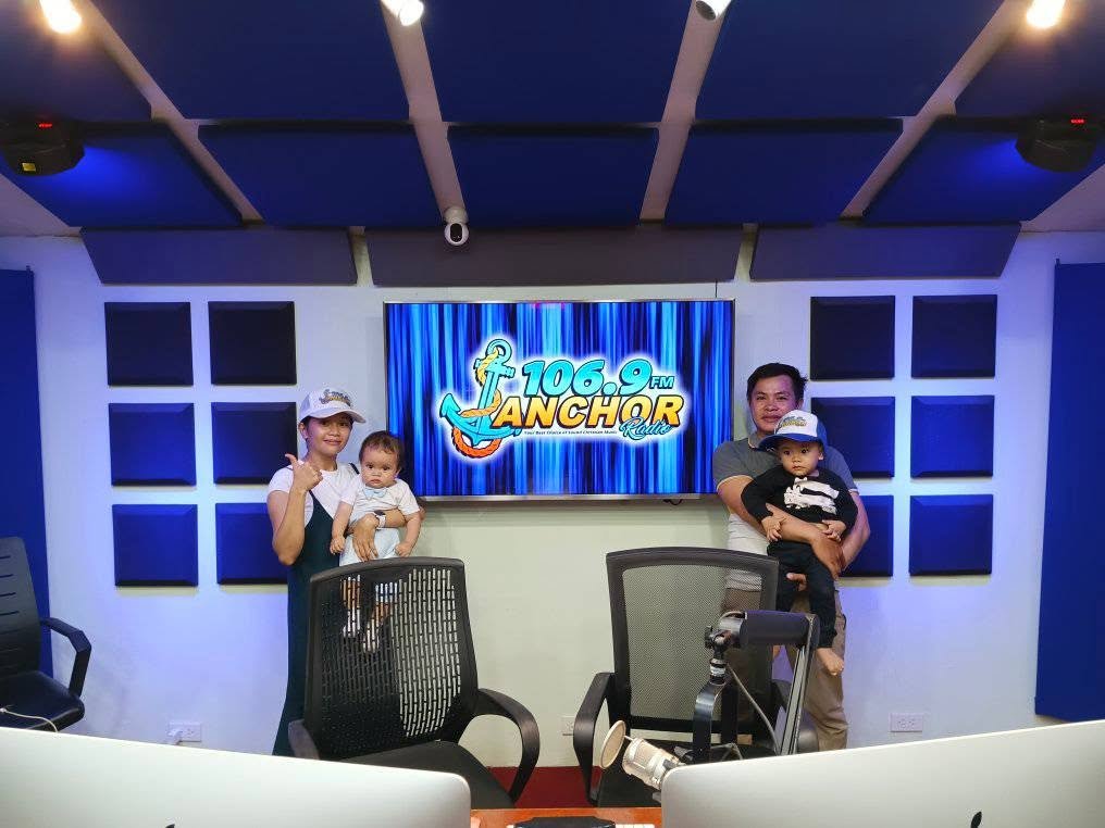 Two women each holding a child, standing in a recording studio with a large screen behind them displaying radio station logo "106.9 FM Anchor Radio." The women and children are smiling, and there are chairs and soundproof panels in the studio.