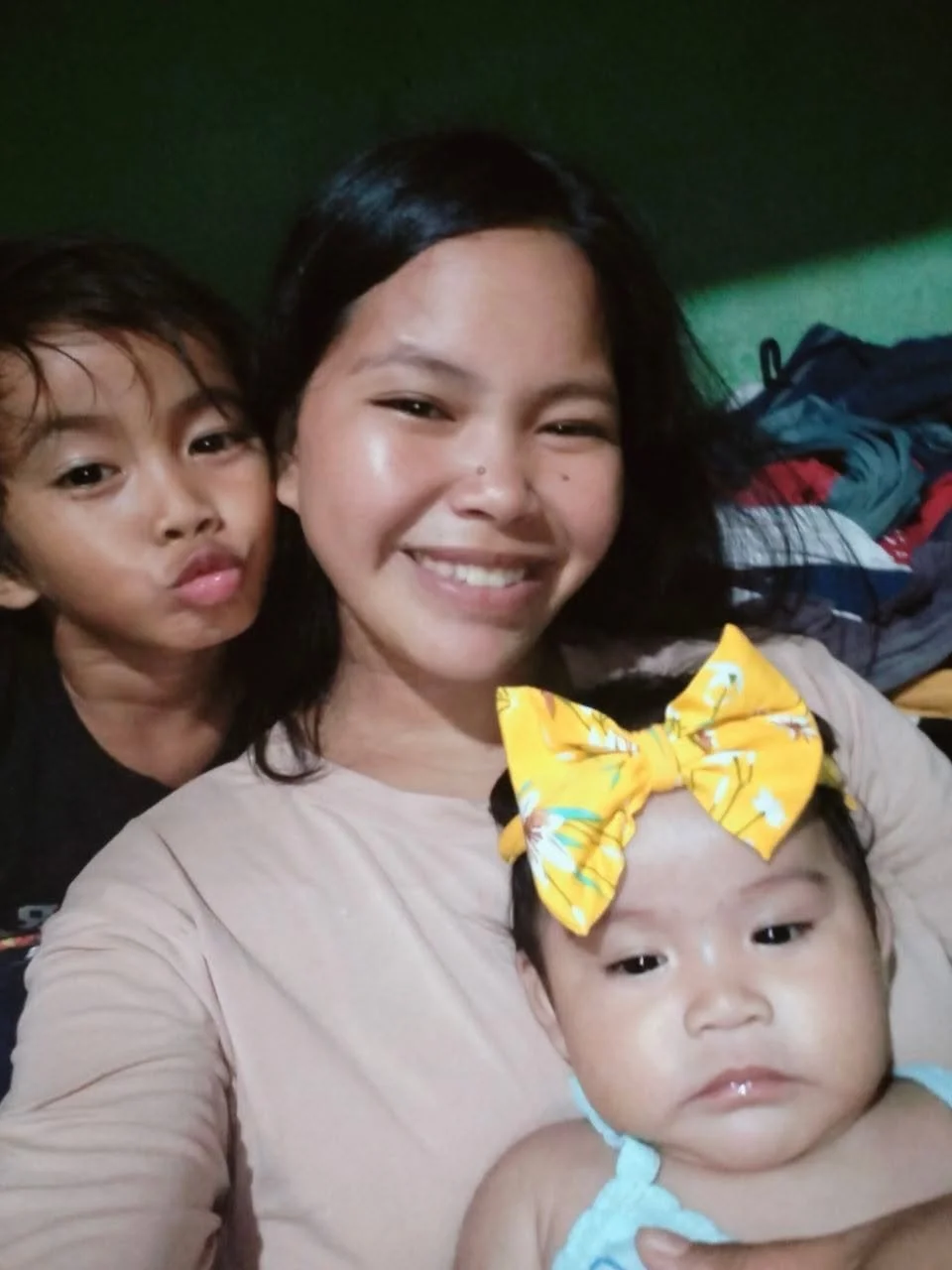 A woman taking a selfie with two children, a boy on her left with wavy hair making a pouty face and a girl with a yellow bow in her hair on her chest, all smiling and close together inside a room.