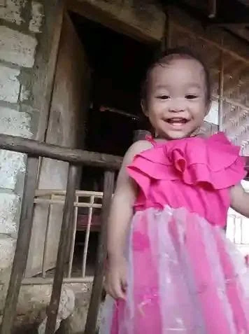A young girl in a pink dress smiling in front of a rustic wooden building.