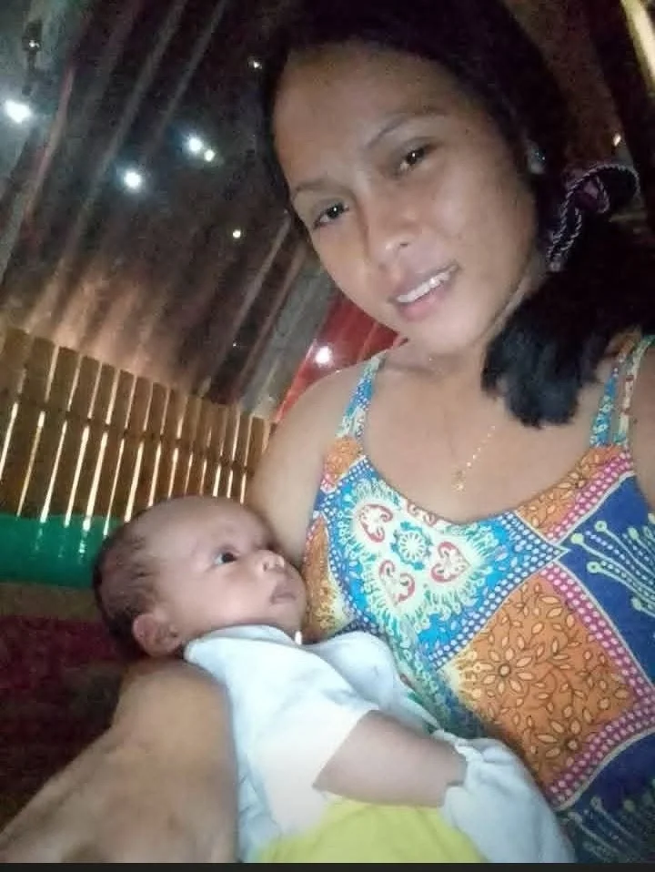 A woman in colorful clothing holding a baby in a room with wooden walls and small windows.