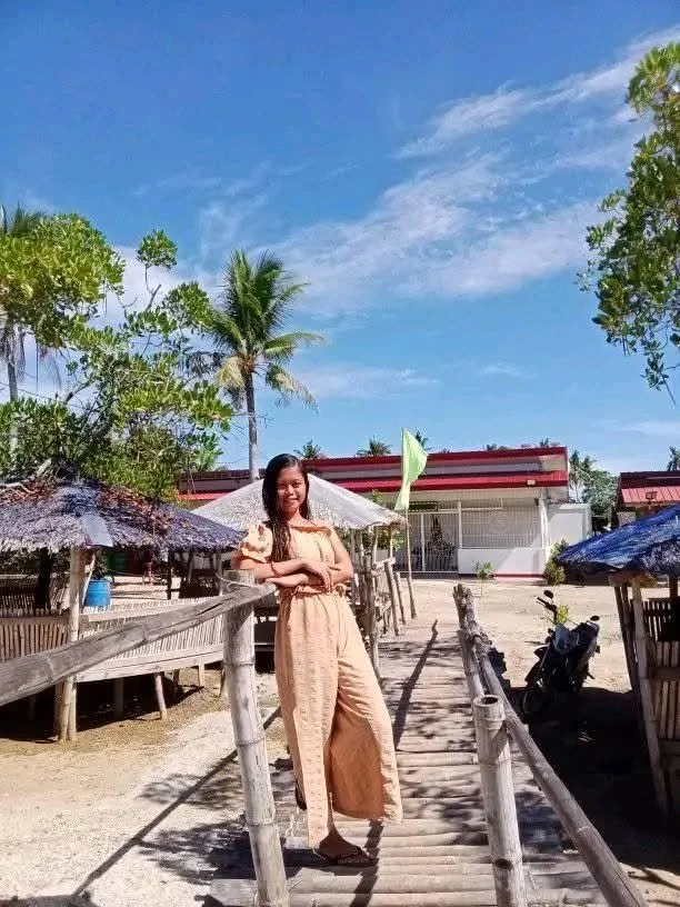 Woman in beige jumpsuit standing on a wooden bridge in a tropical setting with palm trees, huts, and a blue sky.