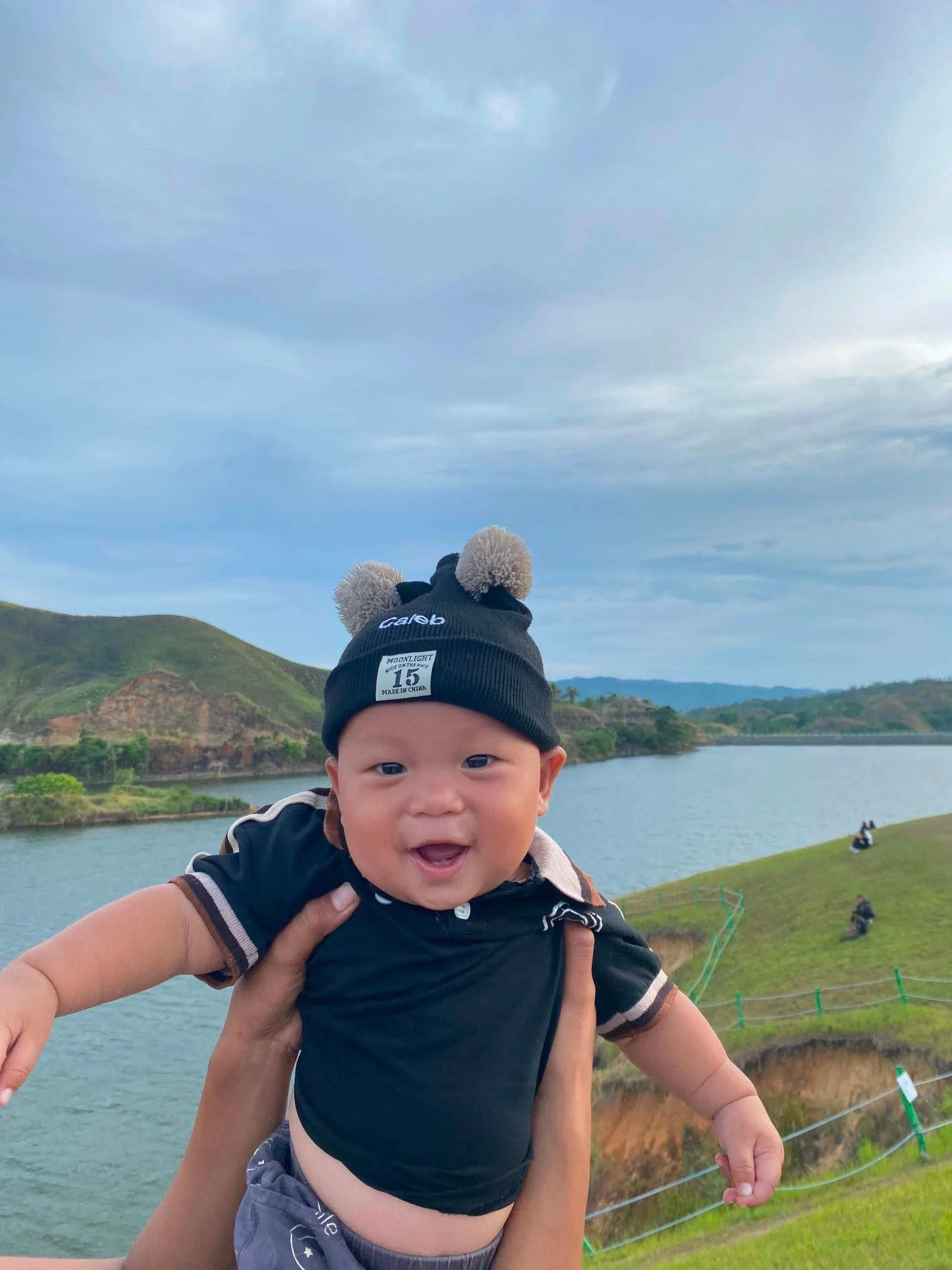 A young child with a baseball cap smiling while being held up by an adult near a lake with green hills and cloudy sky in the background.