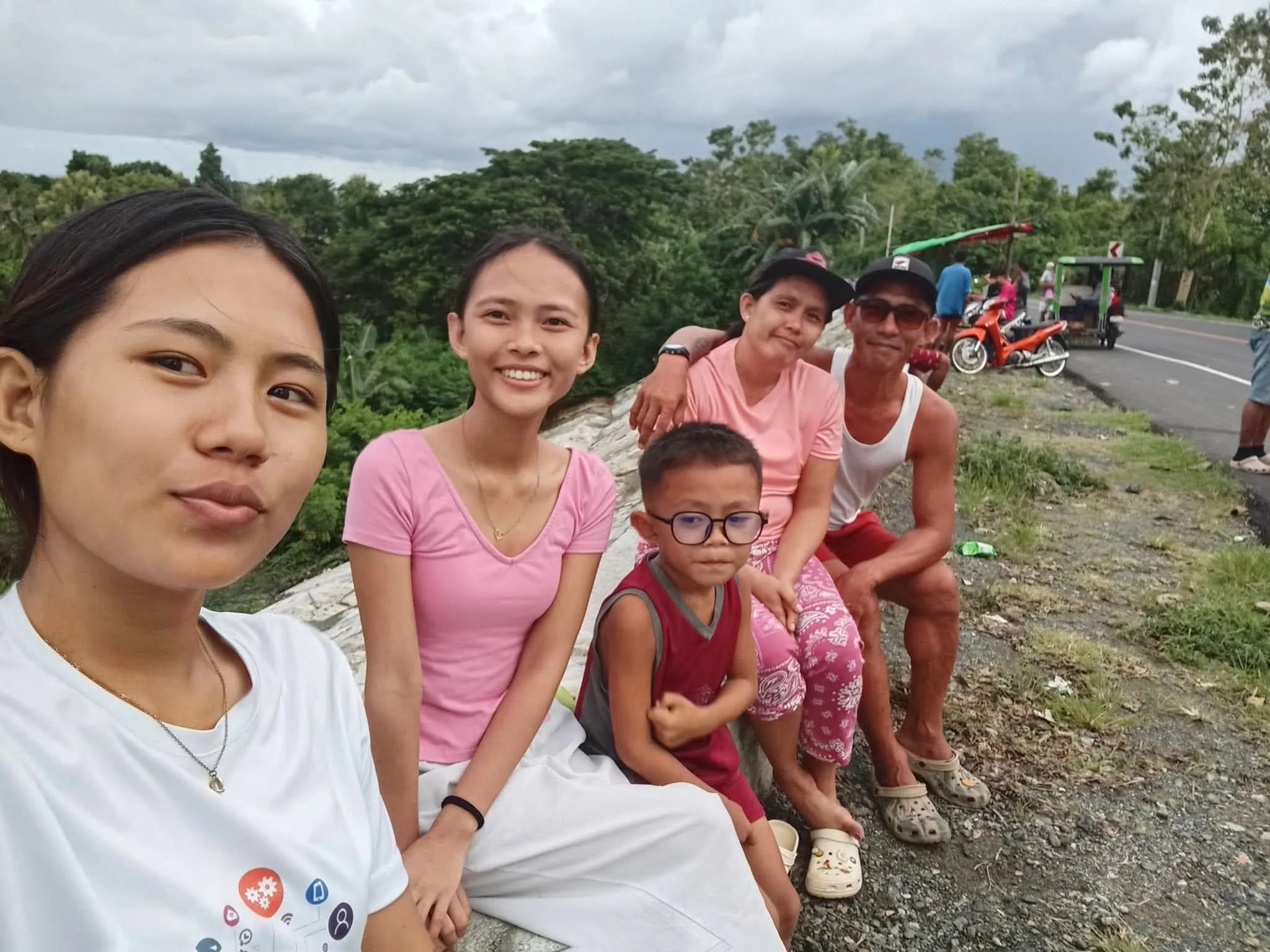A group of diverse people, including children and adults, sitting outdoors on a rural roadside, smiling, with green trees and cloudy sky in the background.