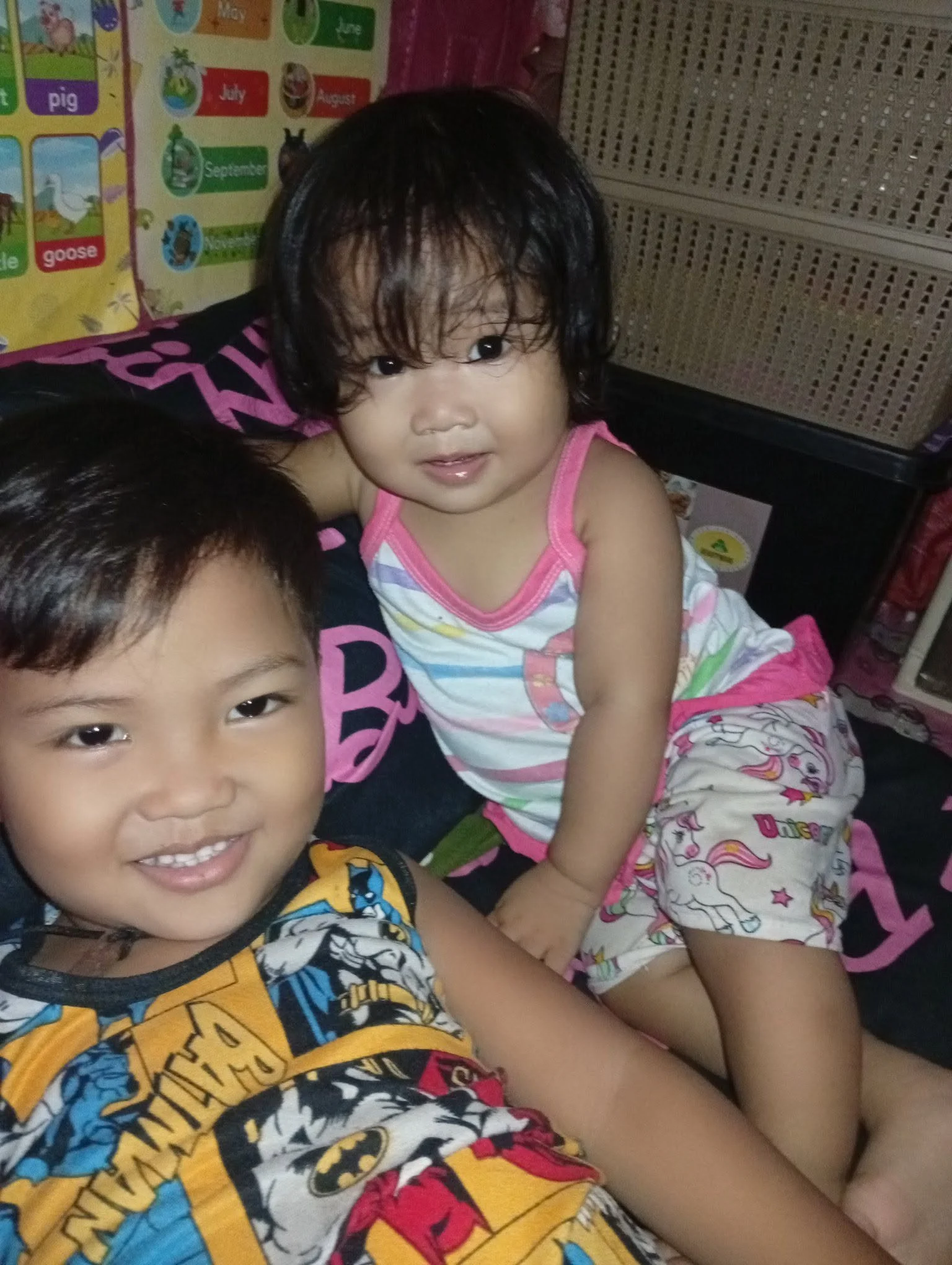 Two young children with dark hair smiling and looking at the camera in a room, with colorful educational posters and decor in the background.